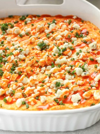 A white baking dish filled with creamy buffalo chicken dip topped with blue cheese, chives, and buffalo sauce.
