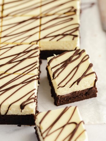 Sliced cheesecake brownies drizzled with melted chocolate on a piece of parchment paper.