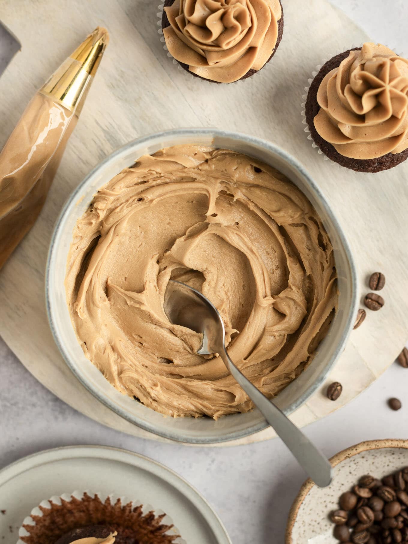 An overhead view of a bowl of coffee buttercream frosting, with two cupcakes. 