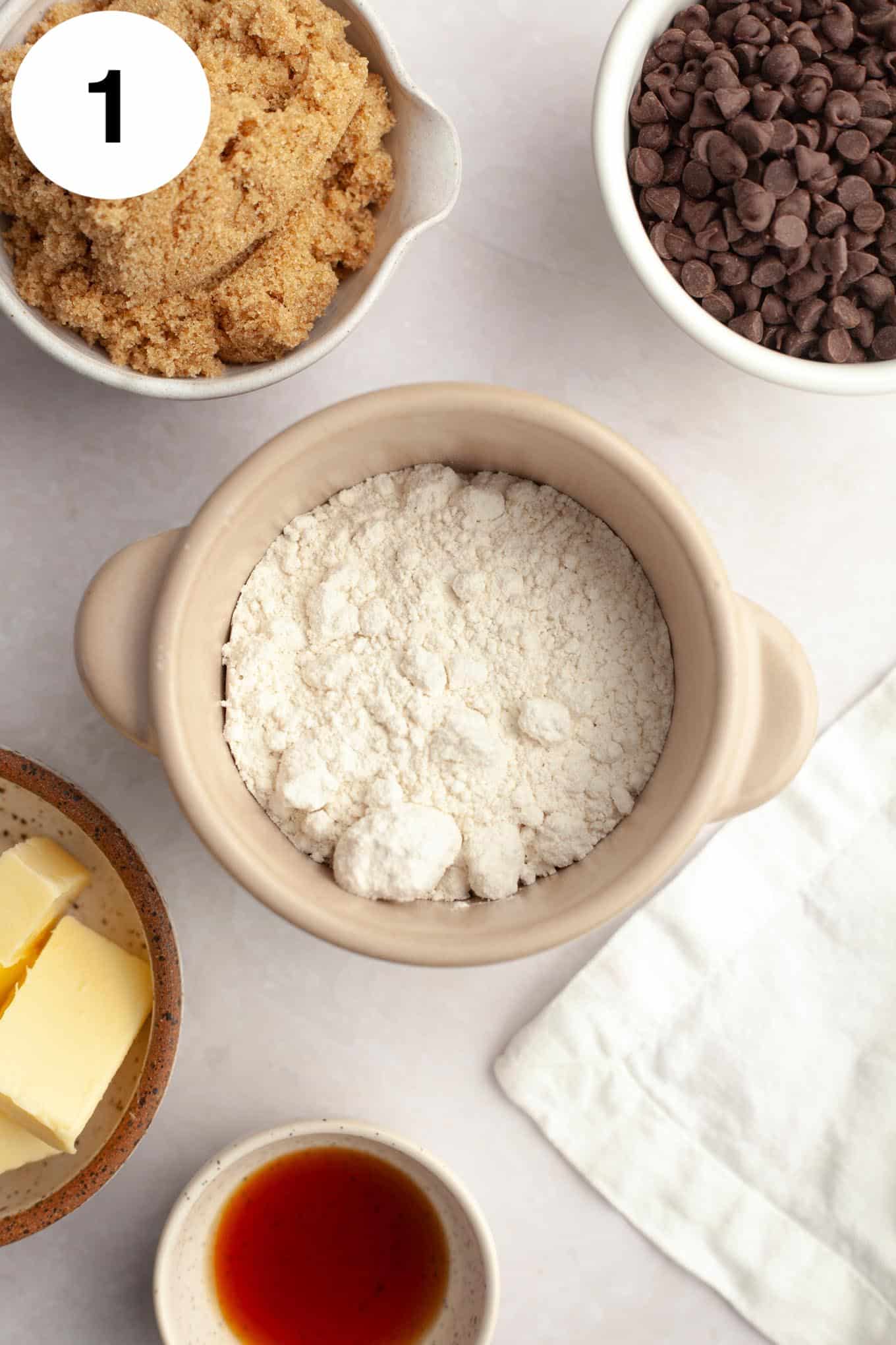 An overhead view of individual bowls of brown sugar, chocolate chips, flour, butter, and vanilla. 
