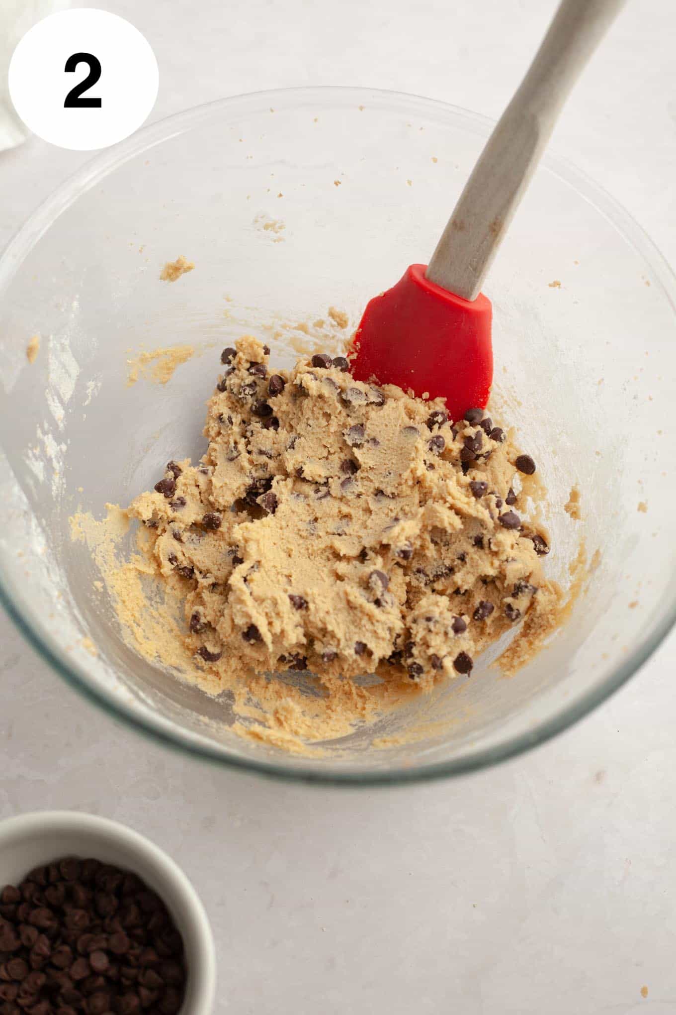 An overhead view of edible chocolate chip cookie dough in a glass mixing bowl, with a rubber spatula. 