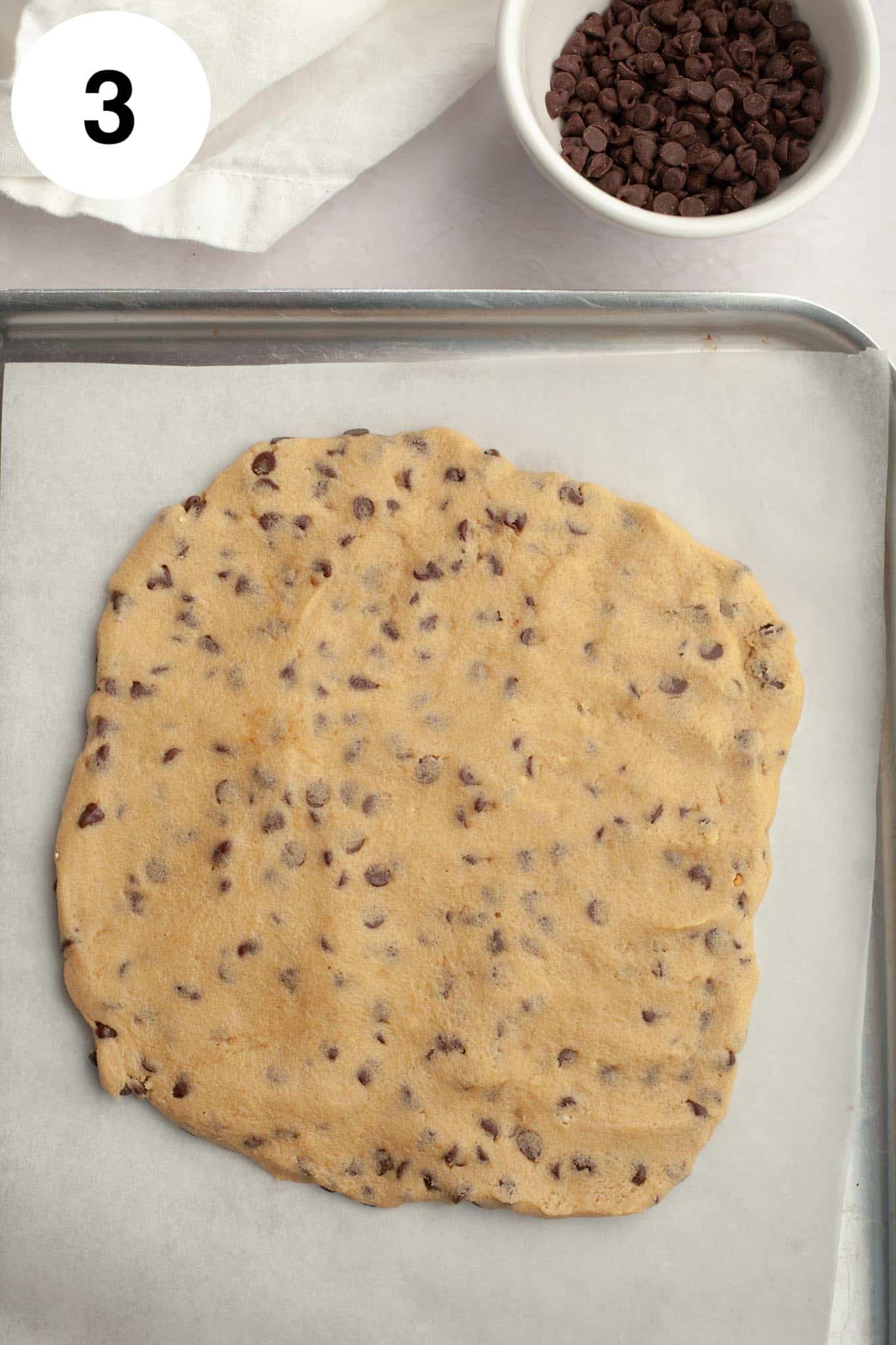 An overhead view of edible chocolate chip cookie dough spread on a parchment paper-lined baking sheet. 
