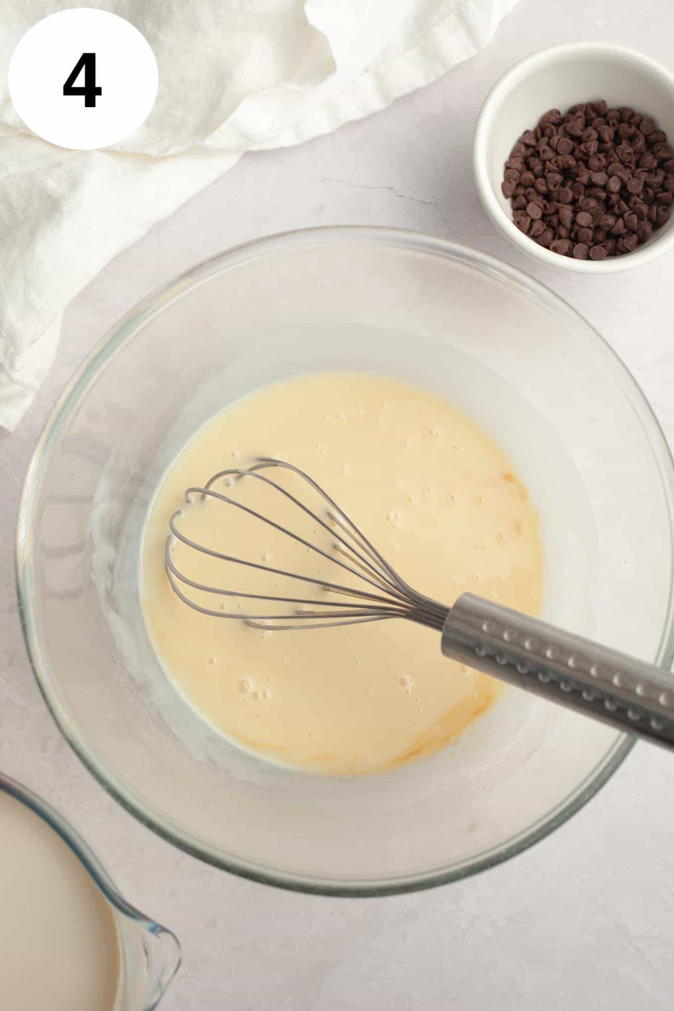 An overhead view of a sweetened condensed milk mixture in a glass mixing bowl, with a whisk