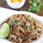 Shredded carnitas meat sitting on a white plate with cilantro and cheese in the background.