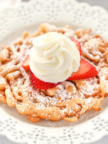 A funnel cake topped with strawberries and homemade whipped cream sitting on a decorative white plate.