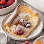 A slice of German pancake dusted with powdered sugar and topped with fresh berries.