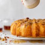A baked rum cake resting on a round cooling rack and the rum glaze being poured over the top.