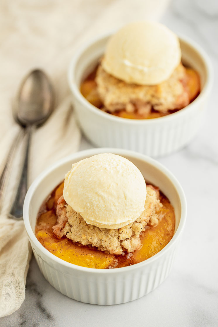 A white ramekin on a marble surface filled with peach cobbler and scoop of ice cream on top.