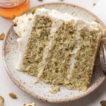 A slice of pistachio cake on a speckled plate. A small jar of honey and pistachios are near the plate.