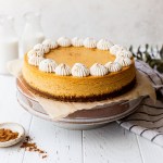 A pumpkin cheesecake on top of a cake stand with milk and cinnamon around it.