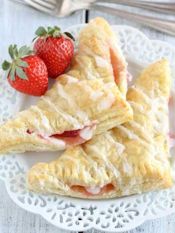 Two strawberry turnovers on a decorative white plate. Two strawberries rest on the plate.