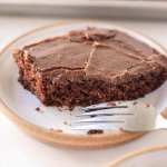 A slice of Texas sheet cake on a white speckled plate. A bite has been taken out of the slice and a fork rests on the side of the plate.